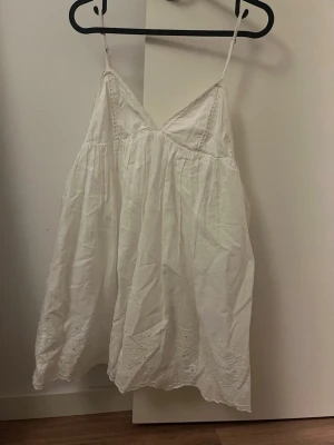 Vit somrig klänning med spetsdetaljer - Luftig vit klänning med tunna axelband och v-ringning. Klänningen har fina spetsdetaljer längs bysten och broderade kanter nertill. Perfekt för varma dagar. Har en liten prick på höger sida men syns knappt.