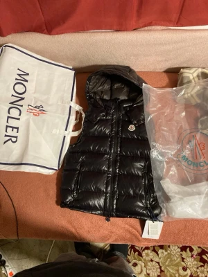 Svart dunväst från Moncler med huva - Snygg svart dunväst från Moncler med glansig finish och avtagbar huva. Västen har två dragkedjefickor framtill och klassisk Moncler-logga på bröstet. Perfekt för lager-på-lager och ger en clean streetwear-look.