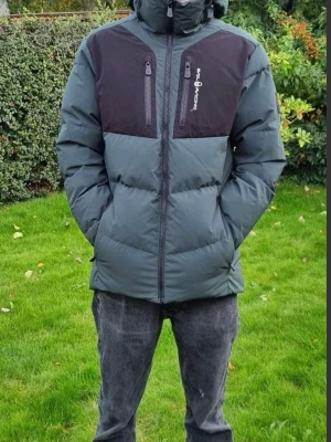 Grå dunjacka från Sail racing - Snygg grå dunjacka från Sail Racing med svarta detaljer på bröst och axlar. Jackan har två vertikala bröstfickor med dragkedja, hög krage och huva. Perfekt för kalla dagar och har en modern, normal passform.