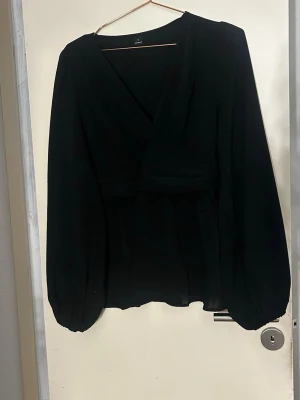 Svart omlottblus med ballongärm - Stilren svart omlottblus med långa ballongärmar och markerad midja. Blusen har v-ringad hals och är tillverkad i ett tunt, lätt material som ger ett snyggt fall. Perfekt att matcha med jeans eller kjol för en trendig look. Använd en gång