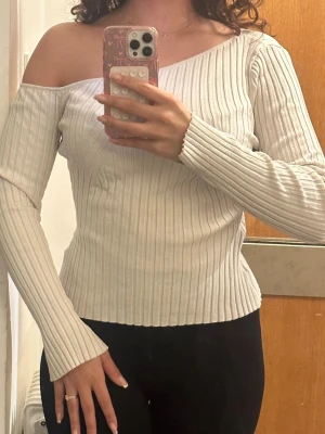 Off shoulder ribbad stickad tröja - Trendig off shoulder stickad tröja i ljusbeige med ribbad struktur. Tröjan har lång ärm och en asymmetrisk halsringning som ger en cool och modern look. Perfekt att styla med jeans eller kjol för en snygg outfit.
