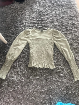 Ljusgrön topp med puffärmar - Trendig ljusgrön topp med puffärmar och smockad kropp. Långa ärmar med smock vid handlederna och volangkant nedtill. Tillverkad i ett mjukt, stretchigt material som ger en snygg passform och cool look.