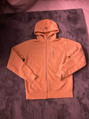 Orange hoodie från Sail Racing - Säljer en orange hoodie från Sail Racing med dragkedja framtill och en bröstficka med zip. Tröjan har broderad logga på bröstet och huva med liten logga. Tillverkad i mjukt material, perfekt för chill dagar. Ribbade muddar vid ärmslut och nederkant.