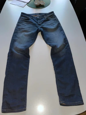 Mörkblå raka jeans från Jack and Jones - Säljer ett par mörkblå jeans från Jack and Jones med raka ben, en snygg wash och snygga slitningar vid knän och lår. Klassisk  femficksmodell med läderpatch bak i midjan. Har unika detaljer som orange fickor och sömmar. Tveka inte på att höra av dig om du undrar något.
