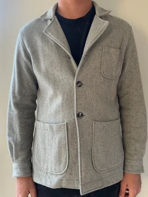 Ljusgrå ullkavaj/overshirt från john henric, storlek S - Stilren ljusgrå kavaj i ullmix med tre utanpåliggande fickor och två knappar framtill. Klassisk krage och enkel design som passar till många olika looks. Perfekt för dig som gillar en avslappnad men ändå uppklädd stil.