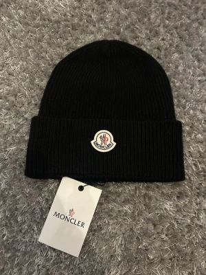 Svart Moncler mössa  - Snygg svart ribbstickad mössa från Moncler med klassisk uppvikt kant och Moncler-logga framtill. Tillverkad i mjuk ull som håller dig varm och ger en clean look. Perfekt accessoar för kalla dagar och enkel att matcha med olika outfits.
