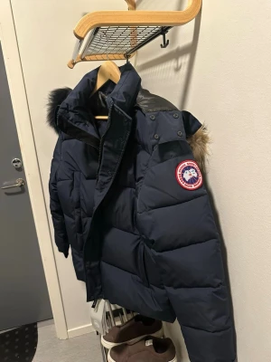 Mörkblå dunjacka från Canada Goose - Snygg mörkblå dunjacka från Canada Goose med avtagbar huva och pälsdetalj. Jackan har det klassiska Canada Goose-märket på ärmen och stora, praktiska fickor. Perfekt för kalla vinterdagar och riktigt varm tack vare dunfyllningen.