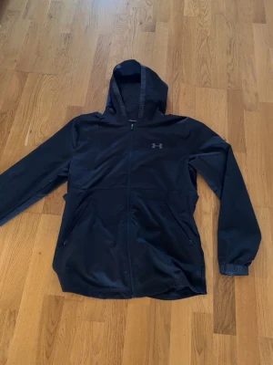 Svart vindjacka från Under Armour - Snygg svart vindjacka från Under Armour med huva och dragkedja framtill. Jackan har två sidofickor med dragkedja, diskret logga på bröstet och detaljer med Under Armour-tryck på huva och ärmslut. Tillverkad i lätt och funktionellt material.