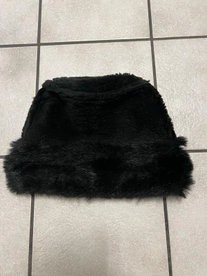 Svart pälsmössa med fluffig kant - Säljer en svart pälsmössa med mjuk och fluffig känsla. Mössan har en bred, glansig pälsimitation runt kanten som ger en lyxig look. Perfekt för kalla dagar när du vill hålla dig varm och snygg.