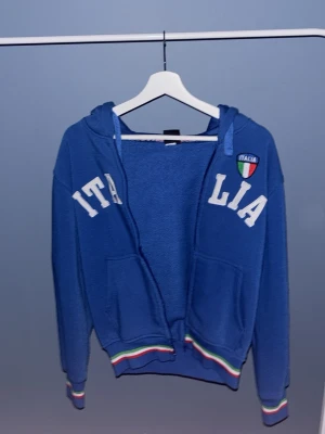 Blå Italia hoodie med huva - Blå hoodie med dragkedja och huva, dekorerad med texten 'ITALIA' i vitt och ett Italienskt märke på bröstet. Färgglada ränder i grönt, vitt och rött vid ärmslut och midja. Två fickor framtill. Perfekt för dig som gillar sportig stil. Tyvär går den inte att knäppa💫