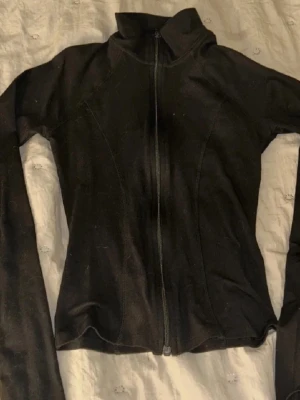 Svart zipjacka med hög krage - Svart jacka med hel dragkedja och hög krage. Jackan har en figurnära passform och långa ärmar, perfekt för en sportig och stilren look. Materialet känns mjukt och stretchigt.