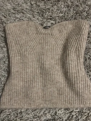 Beige ribbad stickad tubtopp - Säljer en beige ribbad stickad tubtopp med bandeauform. Toppen är ärmlös och har en mjuk, stretchig passform som sitter snyggt på kroppen. Perfekt att styla med jeans eller kjol för en en fest 