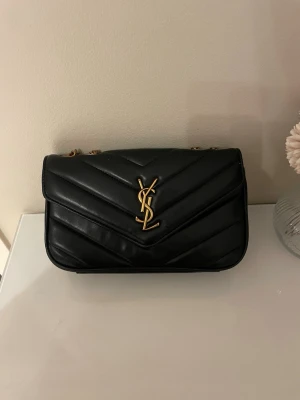 Svart YSL axelväska i läder - Säljer en svart axelväska från Saint Laurent i quiltat läder med guldiga kedjor och ikoniska YSL-loggan framtill. Väskan har en klassisk rektangulär form och elegant design som passar till många olika stilar. Perfekt för dig som gillar lyxiga accessoarer.