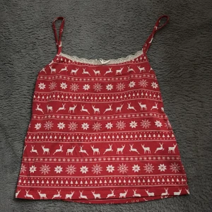 Röd julmönstrat linne med spets - Mysigt rött loungewear-linne med smala axelband och juligt mönster av snöflingor och renar. Dekorerad med vit spetskant upptill och liten rosett i mitten. Perfekt för vinterkvällar och julmys.