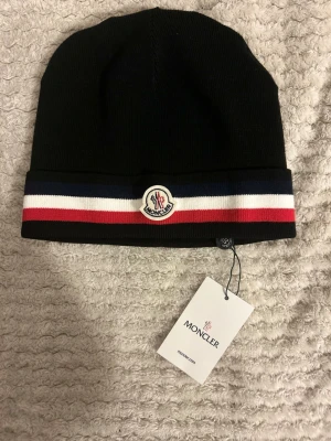 Svart M0ncler mössa med logga - Snygg svart mössa från M0ncler med klassisk M-logga framtill. Mössan har en ribbad kant med ränder i marinblått, vitt och rött. Tillverkad i mjukt stickat material som håller dig varm och stilren under kalla dagar.