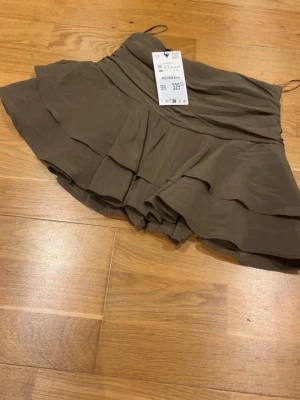 Grön volangkjol med inbyggda shorts i XS - Snygg kort grön/khaki kjol med volanger. Kjolen har en flowy passform och är perfekt för dig som gillar en trendig och avslappnad stil. Tillverkad i ett lätt material som ger ett fint fall. Helt ny med prislapp. 