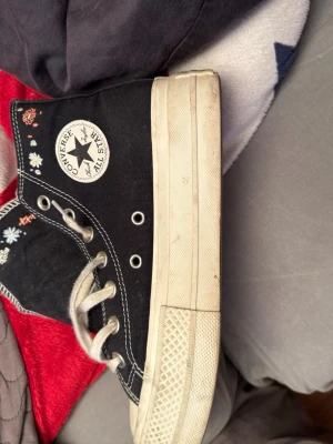 Converse All Star svarta höga sneakers - Svarta Converse All Star sneakers med högt skaft och vita sulor. Skorna har vita snören, metallfärgade öljetter och klassisk logga på sidan. Dekorerade med små broderade blommor i olika färger. Tillverkade i canvas med gummisula.är mer som en 36 i storlek 