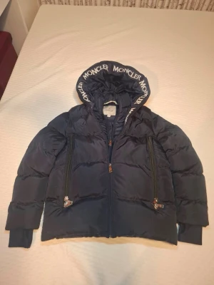 Mörkblå dunjacka från Moncler - Snygg mörkblå dunjacka från Moncler med stor huva där Moncler-loggan är tryckt runt kanten. Jackan har två stora dragkedjefickor framtill och breda muddar vid ärmsluten. Perfekt för kalla vinterdagar och riktigt stilren look.