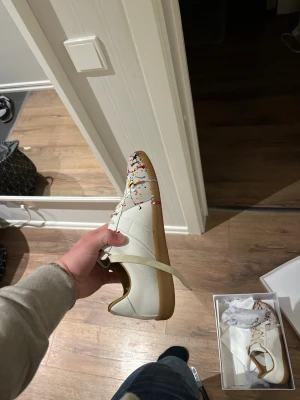 Gats - Snygga vita sneakers med gummisula och coola färgstänk i rött, gult och svart över tån och sidan. Skorna har klassisk låg profil och vita skosnören. Perfekta för dig som vill sticka ut med en unik och kreativ stil.