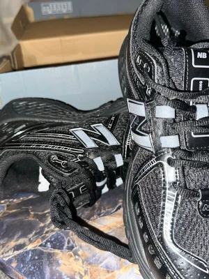 New Balance 1906D svarta sneakers - Svarta New Balance 1906D sneakers med reflekterande detaljer och mesh-paneler för extra ventilation. Snygg design med silvriga inslag och klassisk N-logga på sidan. Perfekta för dig som gillar sportig och trendig stil.