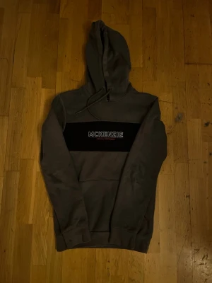 Grå McKenzie hoodie med svart detalj - Säljer en grå hoodie från McKenzie med svart panel över bröstet och vit logga. Klassisk huva med dragsko och stor magficka framtill. Tillverkad i mjukt bomullsmaterial, perfekt för chill dagar. Snygg och enkel design som funkar till det mesta.