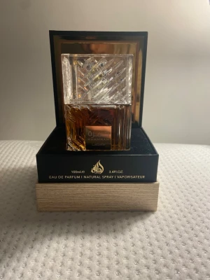 Lattafa Khamrah Eau de Parfum 100ml - Lattafa Khamrah Eau de Parfum kommer i en lyxig, fyrkantig glasflaska med ett elegant mönstrat lock. Flaskan har en varm bärnstensfärg och levereras i en svart och guld ask med exklusiv design. Volymen är 100 ml och parfymen är en naturlig spray.