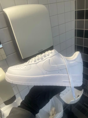 Nike Air Force 1 helvita sneakers - Klassiska Nike Air Force 1 sneakers i helvitt läder med vit sula och vita skosnören. Ikonisk siluett med perforerad tåbox och Nike swoosh på sidan. Perfekta för en clean och stilren look.