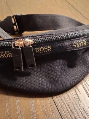 Svart magväska från BOSS nyskick  - Snygg svart magväska från BOSS med justerbart band och guldfärgade dragkedjor. Väskan har tydlig BOSS-logga i guld på framsidan och flera fack med dragkedja. Perfekt att bära crossbody för en trendig look.