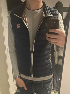 Mörkblå dunväst från Colmar (prisförslag) - Snygg mörkblå dunväst från Colmar med vit dragkedja och Colmar-logga på bröstet. Västen har en quiltad design och två sidofickor med dragkedja. Perfekt att slänga över en hoodie eller tröja för en casual look. En liten defekt är att de är inget handtag på dragkedjan