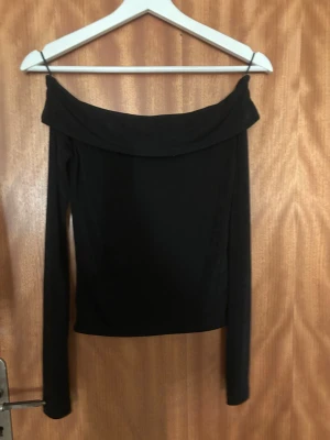 Svart offshoulder topp från Gina tricot - Stilren svart offshoulder topp med långa ärmar och bred krage. Tunn och mjuk i materialet, perfekt att matcha med jeans eller kjol för en trendig look. Enkel och tidlös design som passar till många olika stilar. Super bra skick då den ej är använd många gånger