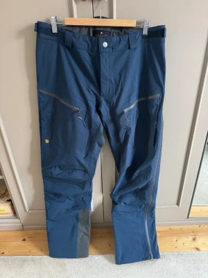 Fjällräven Bergtagen Eco-Shell byxa - 3-lagers skalbyxor som skyddar mot vind och väta på vandringar och skidturer. Vattenavvisande ventilationsdragkedjor längs sidorna, förstärkningar kring hängselpartiet och förstärkningar i benslut. Avtagbara snölås. Passar året runt, både på skidor och till fots. Aldrig använda. Nypris 5999kr. 