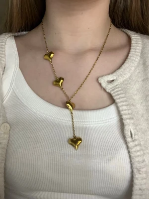 Guldigt halsband med hjärtan - Snyggt halsband i guldfärgad metall med fyra blanka plast hjärtan i rad. Kedjan är tunn och elegant, och hjärtana har en mjukt rundad form som ger ett trendigt intryck. Perfekt accessoar för att lyfta din outfit. Handgjort