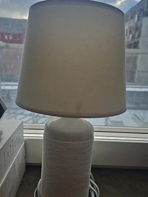 Vit bordslampa med textilskärm - Säljer en stilren vit bordslampa med rund textilskärm och cylinderformad fot i keramik. Lampan har en enkel och modern design som passar bra i de flesta rum. Sladd medföljer.
