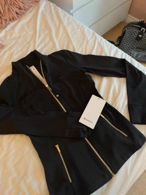Svart jacka från Lululemon med guldiga dragkedjor - Svart jacka från Lululemon med snygga guldiga dragkedjor framtill och på fickorna. Jackan har en figurnära passform och är tillverkad i ett stretchigt syntetmaterial. Perfekt för en sportig och stilren look.