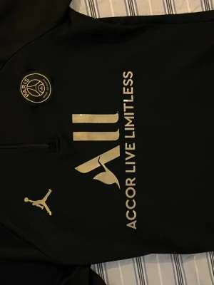Svart PSG träningströja Jordan - Snygg svart träningströja från PSG och Jordan med guldfärgade detaljer, Paris Saint-Germain-logga på bröstet och texten 'Accor Limitless' framtill. Halvlång dragkedja och diskret Paris-tryck på axeln. Perfekt för fotbollsträning eller streetwear.