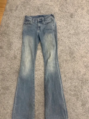 Ljusblå bootcut jeans från true religion - Säljer ett par ljusblå bootcut jeans från True religion med slitningar framtill och klassiska fem fickor. Jeansen har en något utsvängd passform nedtill och detaljerade bakfickor med knapp. Perfekta för en avslappnad och trendig look.