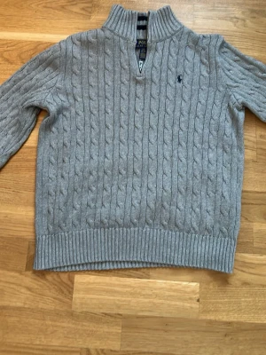 Grå kabelstickad tröja Polo Ralph Lauren - Kabelstickad grå tröja från Polo Ralph Lauren med halv dragkedja och hög krage. Tröjan har ribbstickade muddar och nederkant samt den klassiska broderade loggan på bröstet. Perfekt för lager-på-lager och snygg till jeans.