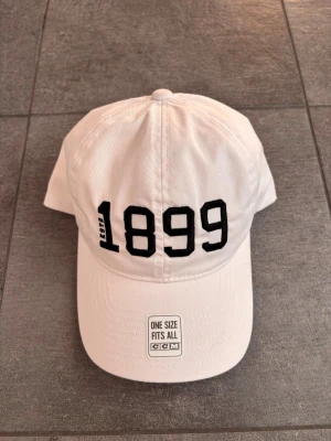 Vit keps från CCM med 1899-tryck - Vit keps från CCM med svart broderat 1899-tryck framtill och justerbar rem baktill. Klassisk basebollmodell i bomullstyg, one size fits all. Perfekt för dig som gillar sportig och stilren look.