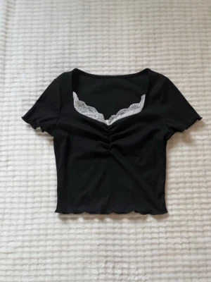 Svart croppad topp med spetsdetalj - Svart croppad topp med korta ärmar och v-ringning dekorerad med vit spets. Toppen har rynk framtill och vågig kant nertill och vid ärmslut. Perfekt för en trendig och ungdomlig stil.