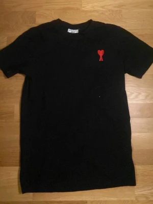 Svart t-shirt från Ami Paris med rött hjärta - Svart t-shirt från Ami Paris med klassisk passform och korta ärmar. På bröstet sitter ett rött broderat hjärta med bokstaven A. Tillverkad i mjuk bomull som känns skön mot huden. Perfekt för en clean och stilren look.