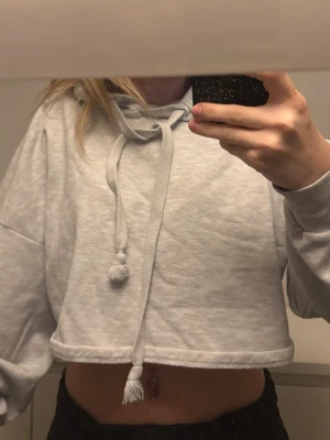 Ljusgrå cropped hoodie med huva - Säljer en ljusgrå cropped hoodie med huva och långa ärmar. Hoodien har dragsko med tofsar och ribbade muddar vid ärmsluten. Perfekt för en avslappnad och trendig look. Materialet känns mjukt och bekvämt.