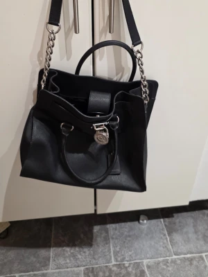 Svart handväska från Michael Kors - Snygg svart handväska från Michael Kors i slätt läder med silverfärgade metalldetaljer och kedjeaxelrem. Väskan har dubbla handtag, dekorativt hänglås med MK-logga och praktisk innerficka. Perfekt för dig som vill ha en stilren och trendig accessoar.