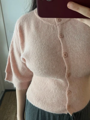 Ljusrosa kofta med kort ärm från Gina Tricot  - Supermjuk ljusrosa kofta med korta ärmar och rund hals. Koftan har knappar framtill och är stickad i ett fluffigt material.