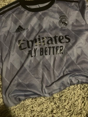 Real Madrid  Adidas  - Snygg lila tröja från Real Madrid med svart mönster och svarta detaljer. Adidas-logga och klubbmärke på bröstet samt Emirates Fly Better-tryck på framsidan. 