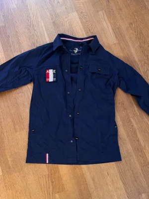 Marinblå skjorta från WHO's A.C.E - Snygg marinblå skjorta från WHO's A.C.E med lång ärm och knappar framtill. Skjortan har patchdetalj på bröstet med text och röd/vit/blå färg samt dekorativ rand på insidan av kragen. Perfekt för en stilren och sportig look.