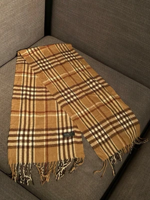 Rutig halsduk från Burberrys - Klassisk rutig halsduk från Burberrys i beige, brunt och vitt. Tillverkad i 100% lammull och har fransar i ändarna. Märkt 'Made in England'. Perfekt accessoar för att lyfta din outfit med en tidlös brittisk stil.