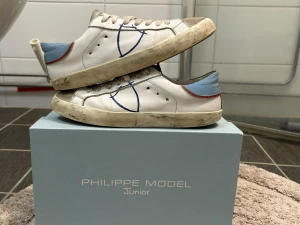 Philippe Model vita sneakers junior - Vita sneakers från Philippe Model Junior med blå och röda detaljer. Skorna har en låg profil, vita snören och en blå logga på sidan. Hälpartiet är ljusblått med en röd linje. Ovandelen är i läder och sulan är vit med slitningar.
