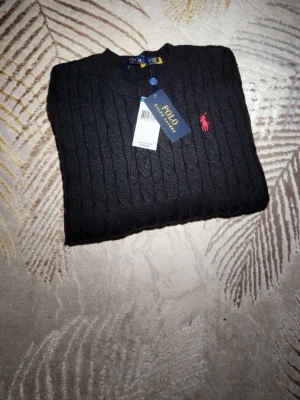 Svart kabelstickad tröja Polo Ralph Lauren - Svart kabelstickad tröja från Polo Ralph Lauren med rund halsringning och klassisk röd broderad logga på bröstet. Tröjan har ribbade muddar och är tillverkad i mjukt material. Perfekt för en stilren och tidlös look.