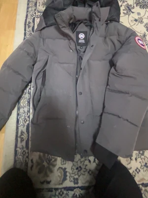 Svart dunjacka från Canada Goose - Säljer en svart dunjacka från Canada Goose med klassisk patch på ärmen och avtagbar huva med fuskpäls. Jackan har dragkedja och knappar framtill, quiltad design och hög krage. Perfekt för kalla vinterdagar och riktigt snygg streetstil, har använt den bara 3gånger den är nästan helt ny 