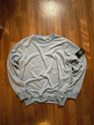 Ljusblå Stone Island sweatshirt - Säljer en ljusblå sweatshirt från Stone Island med klassisk rund hals och ribbade muddar. Tröjan har den ikoniska Stone Island-loggan på vänster ärm. Perfekt för en avslappnad och stilren look.